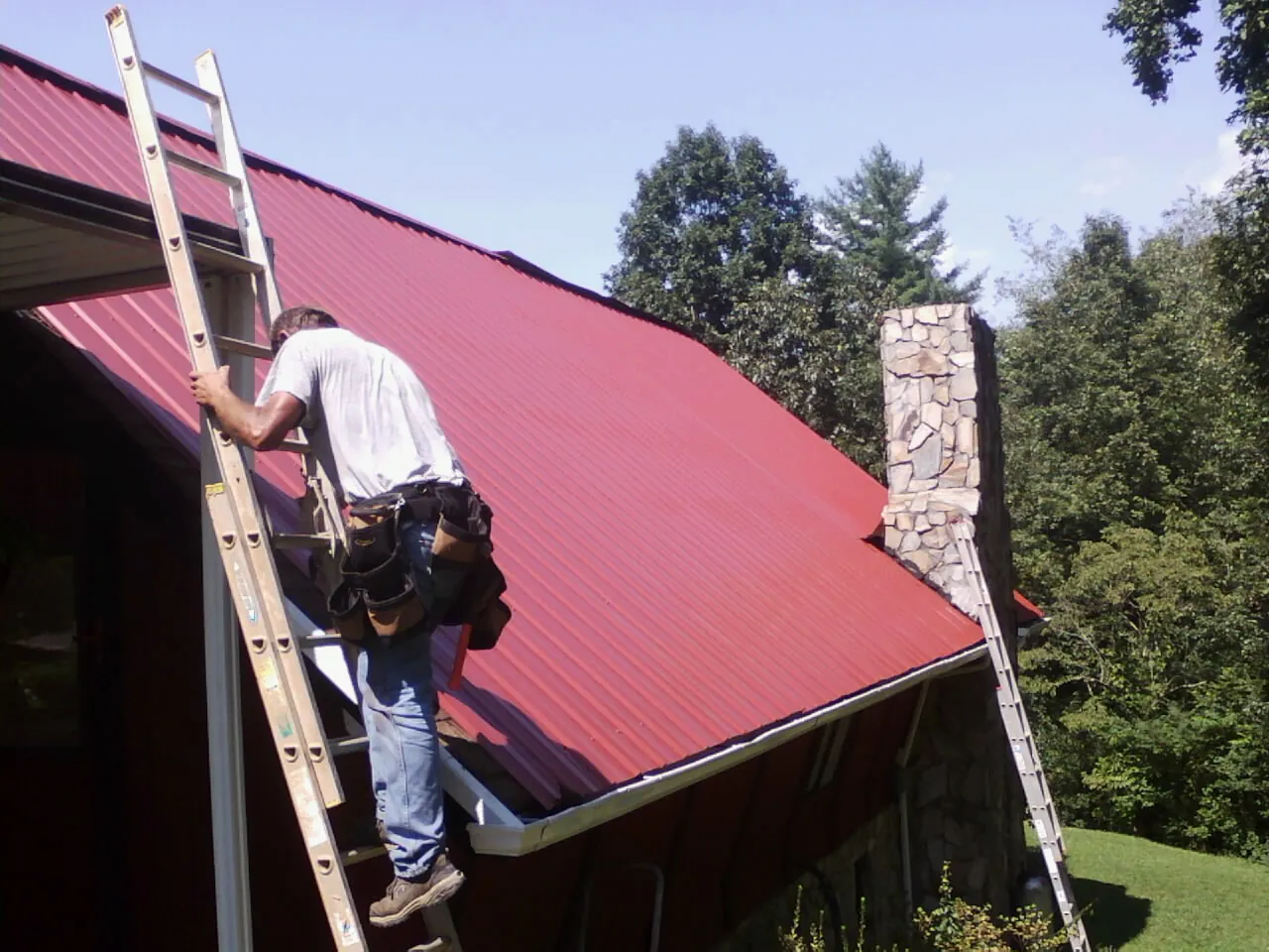 Roofer on a ladder beside a red metal roof and stone chimney — Soffit & Fascia Repair by Roofing Brothers in Shady Cove